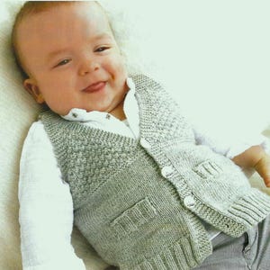 Puede incluir: Un chaleco de bebé de punto gris claro con dos bolsillos delanteros y botones blancos. El chaleco se lleva sobre una camisa blanca con cuello. El bebé lleva pantalones a rayas grises y blancas.