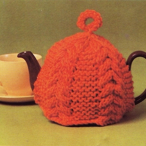 May include: Orange knitted teapot cosy with a cable knit pattern and a loop on top.