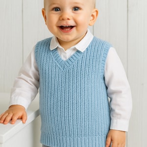Puede incluir: Un chaleco de bebé de punto azul claro con cuello en V, usado sobre una camisa blanca y pantalones grises. El chaleco tiene una textura acanalada. El bebé sonríe, con un fondo de madera blanca.
