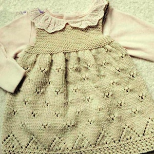 Pode incluir: Um vestido de malha para bebê com gola de renda e uma camisa rosa de manga comprida por baixo. O vestido é bege e tem um padrão delicado.