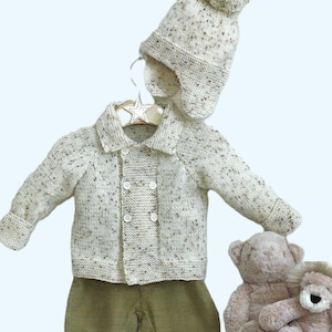 Puede incluir: Un conjunto de bebé tejido en crema con motas marrones, que incluye una chaqueta, un pantalón y un gorro con orejeras y pompón. El conjunto se combina con un pantalón de pana verde oliva. Dos osos de peluche están cerca.