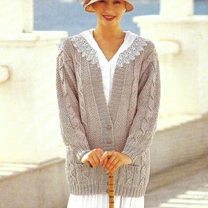 May include: A beige knitted cardigan with a cable knit pattern and a white crocheted collar. The cardigan has a button closure and is worn with a white blouse and a brown hat.