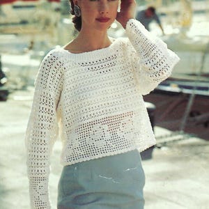 Può includere: Un maglione bianco all'uncinetto a trama aperta con maniche lunghe. Il maglione è abbinato a una gonna azzurra. La donna nell'immagine indossa orecchini e ha i capelli raccolti.