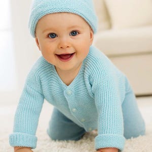 May include: A baby wearing a light blue knitted hat and matching cardigan and pants set. The baby is crawling on a white carpet and smiling. The outfit has a button-up closure.