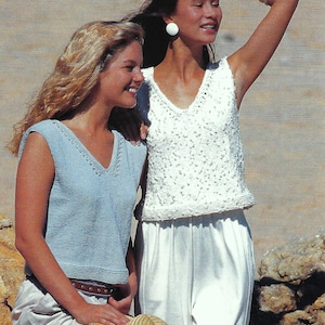 May include: Two women in sleeveless tops. One wears a light blue V-neck top and a brown belt. The other wears a white, textured V-neck top. Both are wearing light-coloured trousers. One woman is holding a straw hat.