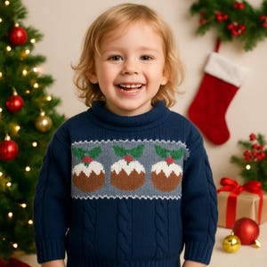 May include: A young child wearing a navy blue Christmas sweater with a cable knit design. The sweater features a pattern of three Christmas puddings with holly and red berries. A Christmas tree and decorations are in the background.