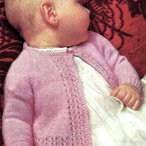 May include: A pink knitted baby cardigan with a delicate lace pattern on the front and sleeves. The cardigan has a button closure and is worn by a baby lying on a red patterned blanket.