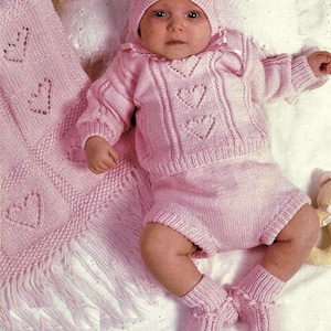 May include: A pink knitted baby outfit with a heart pattern. The outfit includes a hat, sweater, and pants. The baby is wearing matching pink booties.