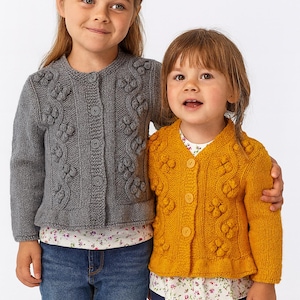 以下が含まれることがあります： 2人の子供がニットカーディガンを着ています。一人は、花柄のデザインのグレーのカーディガンとブルージーンズを着用しています。もう一人は、花柄のデザインのマスタードイエローのカーディガンとネイビーブルーのパンツを着用しています。