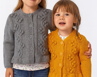 May include: Two young children wearing knitted cardigans. One child is wearing a gray cardigan with a floral design, paired with blue jeans. The other child is wearing a mustard yellow cardigan with a floral design and navy blue pants.