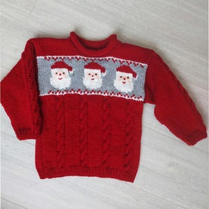 Op de afbeelding: Rode gebreide trui met een kabelbreipatroon en een wit en grijs kerstmanontwerp.
