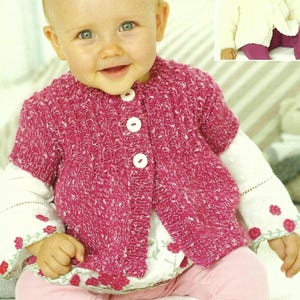 Puede incluir: Un bebé con un cárdigan de punto rosa con botones blancos. El cárdigan tiene un patrón de punto texturizado. El bebé está sentado en una superficie blanca y lleva una camisa blanca con pantalones rosas.