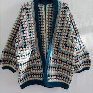 Può includere: Cardigan all'uncinetto fatto a mano, con motivo a granny square. L'indumento aperto presenta quadrati nei toni del verde acqua, crema, lavanda e marrone. Con bordo e maniche verde acqua, appeso a una gruccia nera.