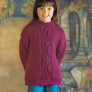 Puede incluir: Una niña pequeña lleva un suéter de punto morado con un patrón de punto de cable. El suéter tiene cuello alto y mangas largas. La niña lleva pantalones de punto azul.