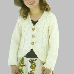 May include: Cream-colored knitted cardigan with heart-shaped wooden buttons. The sweater features cable knit detailing and a scalloped edge. The outfit includes a floral print skirt and a wide-brimmed hat.