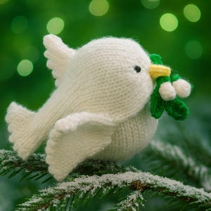 May include: A knitted white dove with a yellow beak and black eyes, holding a sprig of green leaves with white berries. The dove is perched on a snow-covered evergreen branch against a blurred green background.