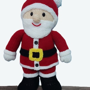 Op de afbeelding: Een gebreide kerstmanpop met een rood pak, muts en handschoenen, een witte baard en bies en zwarte laarzen. De pop heeft een zwarte riem met een gele gesp en een lachend gezicht. Een feestelijke kerstversiering.
