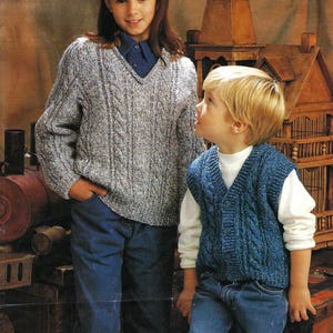 Puede incluir: Dos niños con suéteres de punto. La niña lleva un suéter gris de punto de cable con cuello en V y el niño lleva un chaleco azul de punto de cable con una camisa blanca debajo.