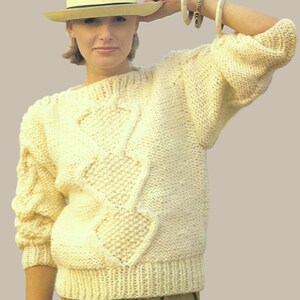 May include: A cream-coloured knitted jumper with a textured diamond pattern. The jumper has a crew neck and long sleeves. A straw hat with a black band is also visible.
