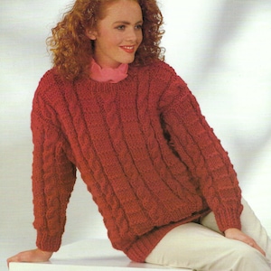 May include: A red cable-knit jumper with a ribbed texture. The jumper has long sleeves and a crew neck. The model is wearing a pink collar underneath the jumper and white trousers. The jumper is a warm, cosy garment.