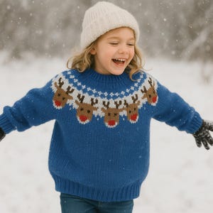 May include: A child wearing a blue knit sweater with a reindeer design and a white beanie hat. The sweater features brown reindeer heads with red noses and a white and brown pattern around the neckline. The child is smiling in a snowy outdoor setting.