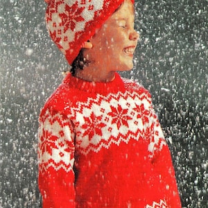 Op de afbeelding: Een kind draagt een rood-witte gebreide trui met een sneeuwvlokkenpatroon en een bijpassende rood-witte gebreide muts met een pompon. Het kind staat in de sneeuw.