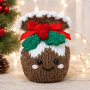 May include: A brown and white knitted pouch shaped like a Christmas pudding with a smiling face, black bead eyes, and pink cheeks. It features a red satin ribbon bow, two green crocheted holly leaves, and three small red crocheted berries on top.