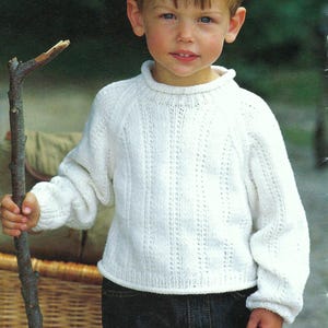 May include: A young boy wearing a white knitted sweater with a cable knit pattern and blue jeans. He is holding a stick in his right hand.