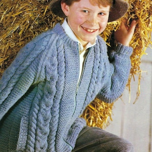 May include: A blue knitted cardigan with a zipper closure and cable knit detail. The cardigan is worn by a young person standing in front of a bale of hay.