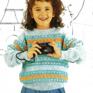 Puede incluir: Un suéter de punto azul claro con rayas naranja y turquesa y un patrón de punto de cable. El suéter tiene un cuello redondo y mangas largas. Una niña lleva el suéter y sostiene una cámara.