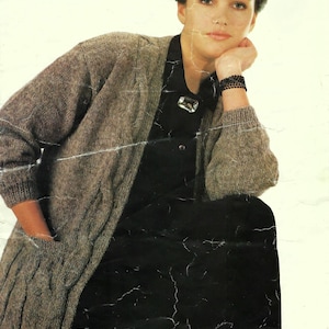 May include: A long-sleeved, open-front knit cardigan in a heathered brown tone featuring cable-knit detailing and two front pockets. The cardigan is worn over a solid black collared shirt adorned with a single clear rectangular brooch at the neckline.