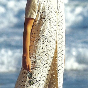 May include: A cream-coloured, open-front crochet vest with a floral pattern. The vest is worn over a short-sleeved shirt. The person is holding sunglasses. The background is a blurred view of the ocean.
