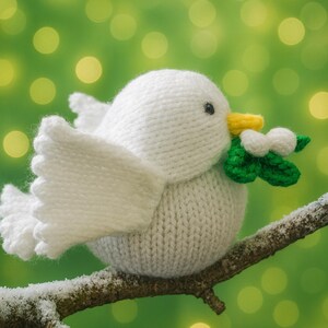 May include: A knitted white bird with outstretched wings perches on a snow-covered branch. The bird has a yellow beak and is holding green leaves with white berries. The background is a blurred green with yellow bokeh.