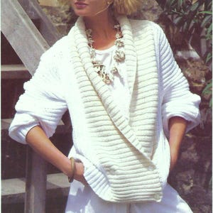 May include: A white knitted cardigan with a wide, folded collar and a white t-shirt underneath. The cardigan has a ribbed texture and is worn with white shorts.