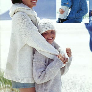 May include: A woman and a child wearing knitted sweaters and hats. The woman is wearing a white sweater with a hood and a black cowboy hat. The child is wearing a light gray sweater and a white beanie. They are standing on a white sandy beach.