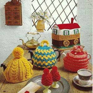 Puede incluir: Una colección de calienta-teteras de punto en varios colores y estilos, incluyendo un calienta-tetera de punto amarillo en forma de sombrero, un calienta-tetera geométrico azul y amarillo, un calienta-tetera a rayas rojo y blanco y un calienta-tetera en forma de casa rojo y blanco. También hay dos calienta-huevos de punto rojo en una bandeja plateada.