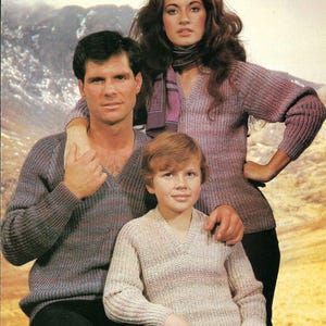 May include: A family portrait featuring three people wearing knitted sweaters. The woman is wearing a purple sweater with a scarf, the man is wearing a blue and purple sweater, and the child is wearing a beige and white sweater.