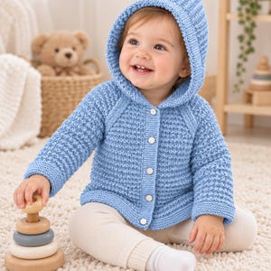 May include: A light blue knitted hooded cardigan featuring a textured waffle stitch pattern, long sleeves, and a front button closure with five silver-toned snaps. The garment is worn over cream-coloured ribbed leggings, providing a soft and cosy look.