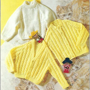 以下が含まれることがあります： ケーブル編みのパターンが施された、白と黄色のニットセーター2枚。セーターは猫柄の背景の上に置かれています。