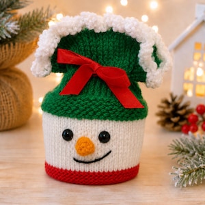 May include: A knitted snowman-themed gift bag featuring a white base with a red bottom edge, a green top section, and a white ruffled trim. The face includes black bead eyes, an orange nose, and a black stitched smile, finished with a red ribbon.