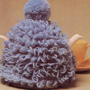 May include: A blue crocheted teapot cozy with a pom-pom on top. The cozy is covering a white teapot with a spout.