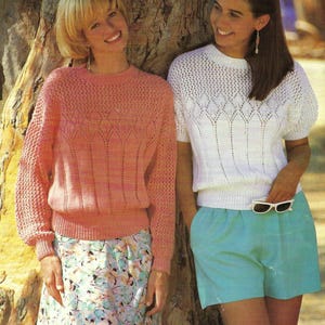 May include: Two women wearing knitted tops and shorts. One woman wears a light pink sweater with a floral skirt. The other woman wears a white short-sleeved top and turquoise shorts. Both are smiling and leaning against a tree.
