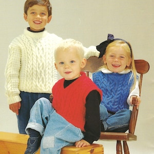 May include: Three children in cable-knit sweaters and vests. A boy in a cream turtleneck sweater stands behind two children. A boy in a red vest sits with a girl in a blue vest on a wooden chair. All are wearing jeans.