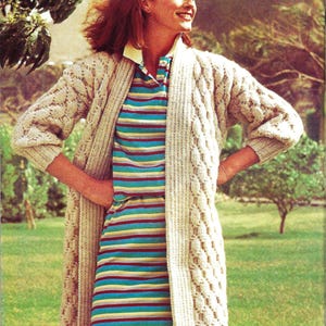 May include: A woman wearing a striped dress with a collared neckline and a long, cream-colored cable-knit cardigan. She is also wearing a red headband. The background is a green lawn with trees.