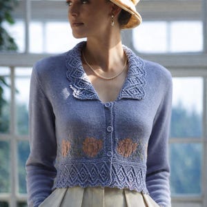 Puede incluir: Un cárdigan azul claro con un diseño floral y un cuello decorativo. El cárdigan está abotonado y se usa con un sombrero y pantalones beige. El diseño floral es en tonos naranja y verde.