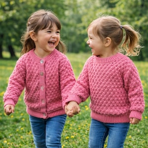 May include: Two young children wearing matching pink knit sweaters and blue jeans, holding hands and laughing. The sweaters have a textured pattern and button closures. The background is a blurred green outdoor setting.