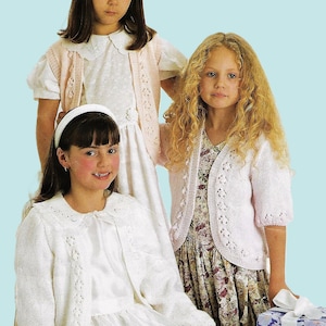 May include: Three young girls in knitted cardigans and dresses. The cardigans are white and pale pink, with decorative openwork patterns. One girl wears a white headband. A gift box with a floral design is visible.