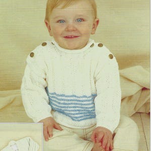 Può includere: Un bambino indossa un maglione bianco lavorato a maglia con strisce orizzontali blu e bottoni decorativi sulle spalle. Il bambino indossa anche pantaloni colorati chiari. Una piccola immagine di un cardigan abbinato si trova nell'angolo in basso a sinistra.