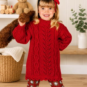 May include: A bright red long-sleeve jumper dress featuring a vertical cable knit pattern and a scalloped hemline, paired with red, white, and black plaid leggings. The jumper has a mock neck collar and a textured knit design throughout.