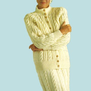 May include: A white cable knit sweater and skirt set with a high neckline and button closure. The set is worn with a white hat.